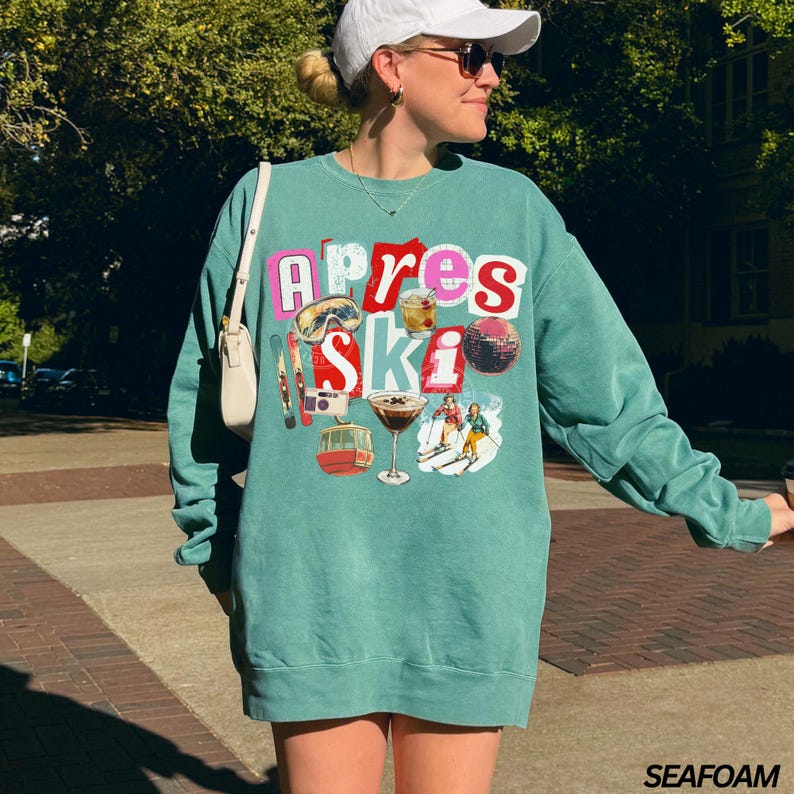 May include: Teal-colored sweatshirt with the words "Apres Ski" in a colorful, layered design. The graphic includes ski equipment, cocktails, and skiers. The sweatshirt is a relaxed fit.