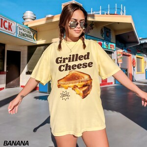 May include: A yellow t-shirt with a graphic of a grilled cheese sandwich and the text "Grilled Cheese" in brown letters. The sandwich has a starburst around it with the text "Extra Cheesy" in smaller brown letters.