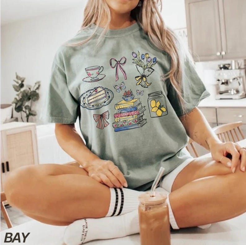 May include: Sage green t-shirt with a whimsical graphic featuring a teacup, cake, books, flowers, bows, butterflies, and a jar of lemons. The word "BAY" is printed at the bottom left corner of the image.