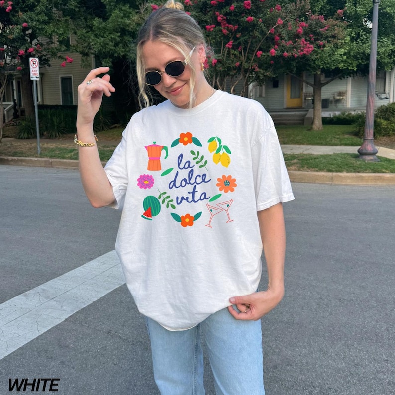 Pu&ograve; includere: Maglietta bianca con un design grafico colorato che presenta limoni, fiori, una caffettiera, anguria e bicchieri da martini. Il testo "la dolce vita" &egrave; stampato al centro del design.