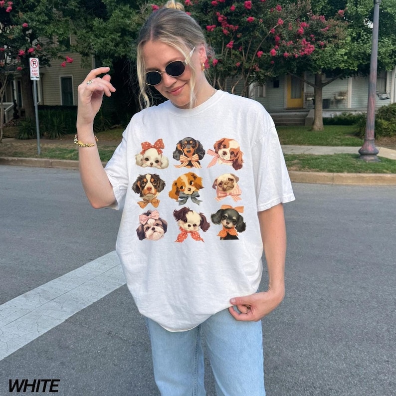 May include: A white t-shirt with a graphic of nine dogs wearing bows. The dogs are all different breeds and have different colored bows. The shirt is being worn by a person with brown hair and sunglasses.
