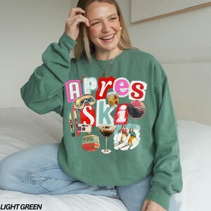 May include: Light green sweatshirt with a colorful graphic that says "Apres Ski." The graphic includes images of ski goggles, cocktails, a disco ball, skis, a camera, and a ski lift. The sweatshirt is a relaxed fit.