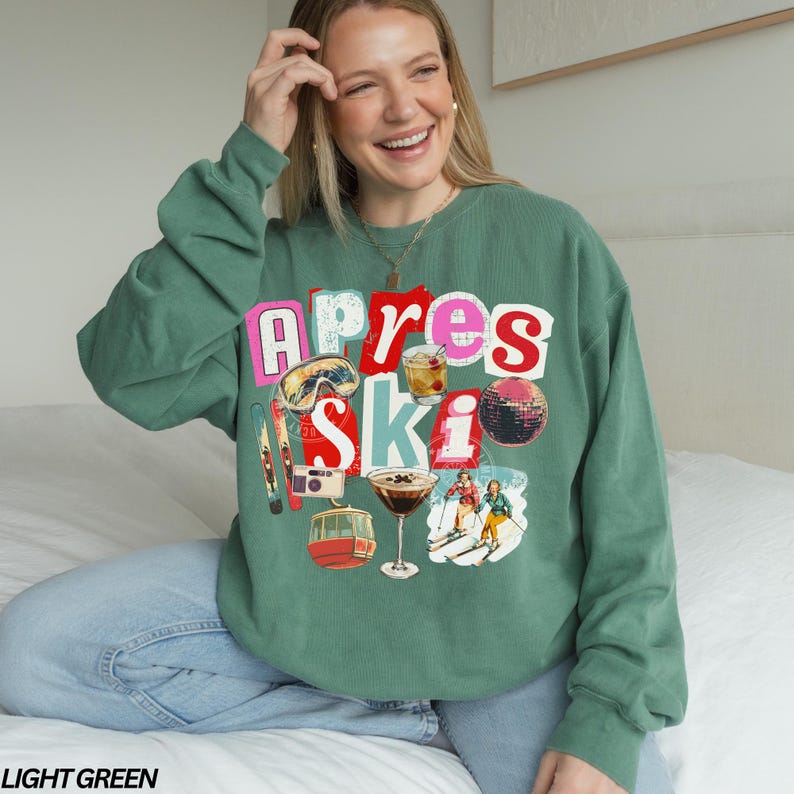May include: Light green sweatshirt with a colorful graphic that says "Apres Ski." The graphic includes images of ski goggles, cocktails, a disco ball, skis, a camera, and a ski lift. The sweatshirt is a relaxed fit.