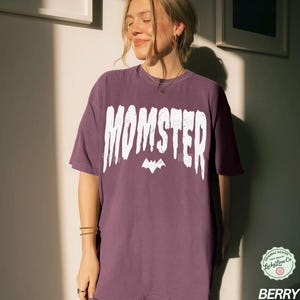 May include: Berry-colored t-shirt with the word "MOMSTER" in white, stylized text above a bat graphic. The shirt has a relaxed fit and short sleeves. The design includes a small logo in the lower right corner.