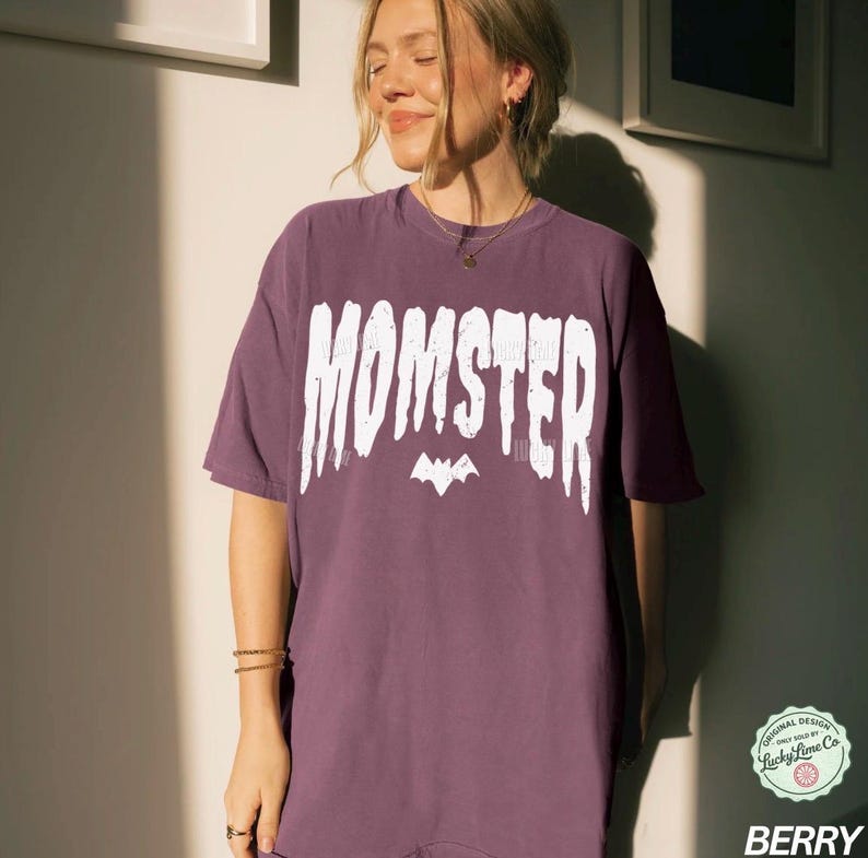 May include: Berry-colored t-shirt with the word "MOMSTER" in white, stylized text above a bat graphic. The shirt has a relaxed fit and short sleeves. The design includes a small logo in the lower right corner.