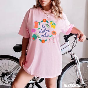 Pu&ograve; includere: Una maglietta rosa oversize con una stampa grafica di un cerchio con il testo "la dolce vita" in blu e illustrazioni colorate di una caffettiera, un'anguria, limoni, fiori e bicchieri da martini.
