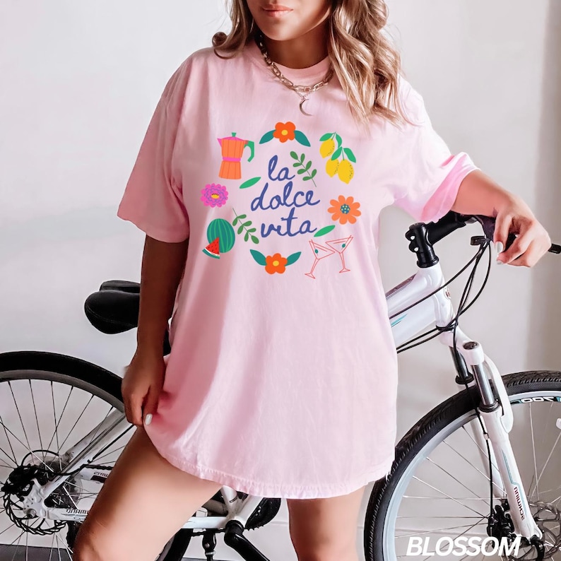 Pu&ograve; includere: Una maglietta rosa oversize con una stampa grafica di un cerchio con il testo "la dolce vita" in blu e illustrazioni colorate di una caffettiera, un'anguria, limoni, fiori e bicchieri da martini.