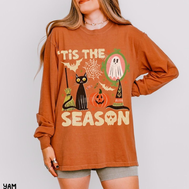 Może przedstawiać: Koszulka z długim rękawem w kolorze rdzawym z okrągłym dekoltem i nadrukiem graficznym z napisem 'TIS THE SEASON'. Wz&oacute;r zawiera czarnego kota, miotłę, pajęczynę, ducha w zielonej ramce, dynię, kapelusz czarownicy oraz czaszkę.