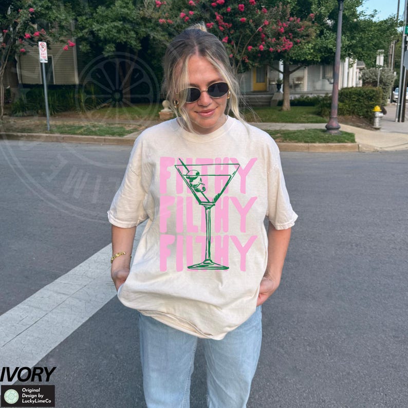 Pode incluir: Uma camiseta branca com um gr&aacute;fico de copo de martini rosa e o texto "Filthy Filthy Filthy" impresso na frente.