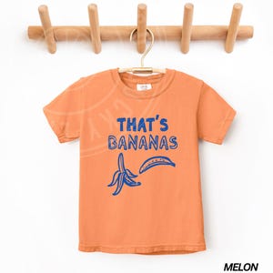 Op de afbeelding: Oranje T-shirt met korte mouwen met een blauwe afbeelding van een banaan en de tekst "That's Bananas".