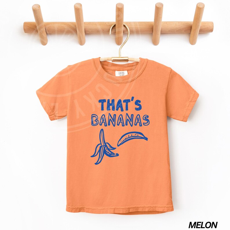 Op de afbeelding: Oranje T-shirt met korte mouwen met een blauwe afbeelding van een banaan en de tekst "That's Bananas".