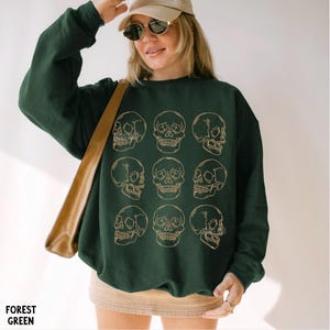 May include: A forest green long-sleeve sweatshirt featuring a grid of nine gold-colored line art skull illustrations. The crewneck garment has a relaxed fit and is worn with a tan baseball cap and a tan skirt, with a tan shoulder bag strap visible.