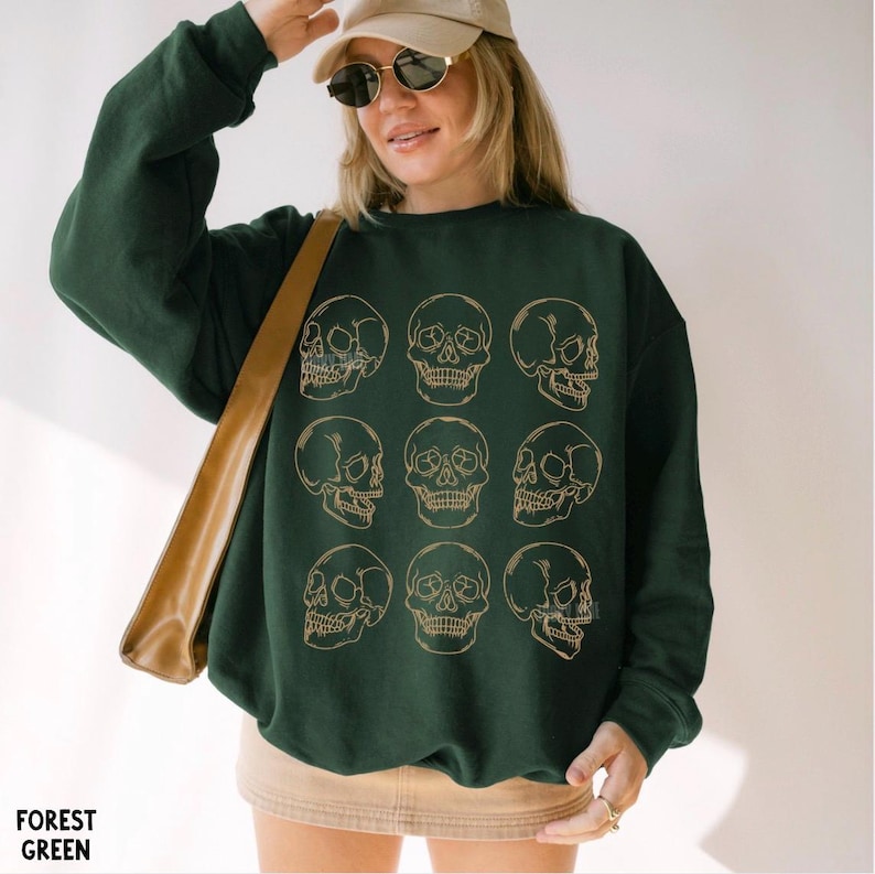 May include: A forest green long-sleeve sweatshirt featuring a grid of nine gold-colored line art skull illustrations. The crewneck garment has a relaxed fit and is worn with a tan baseball cap and a tan skirt, with a tan shoulder bag strap visible.
