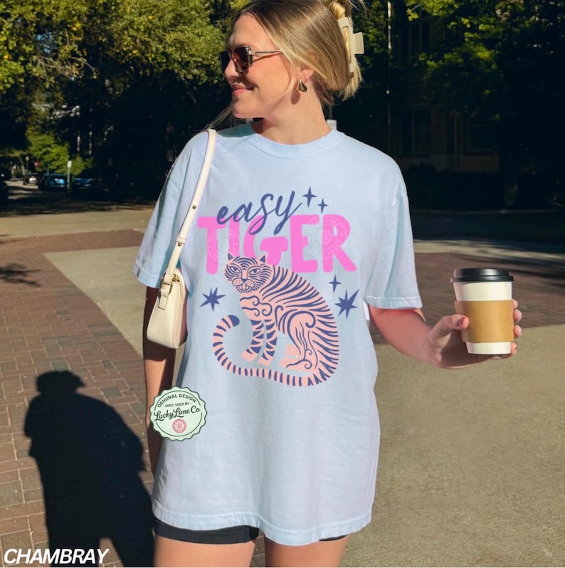 Puede incluir: Camiseta azul claro con la frase "easy TIGER" en rosa y un gr&aacute;fico de tigre rosa y azul. Una persona lleva la camiseta y sostiene una taza de caf&eacute;.