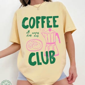 Peut inclure: Un t-shirt jaune p&acirc;le &agrave; manches courtes avec le texte vert COFFEE CLUB, le texte manuscrit vert all every day day, et une illustration au trait rose repr&eacute;sentant une cafeti&egrave;re italienne et une assiette avec un croissant.