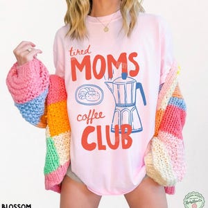 May include: A light pink t-shirt featuring the red text "tired MOMS" and "coffee CLUB" in a bold font, with a blue line-art illustration of a moka pot and a donut on a plate. The shirt is worn under a chunky, multi-colored knit cardigan.
