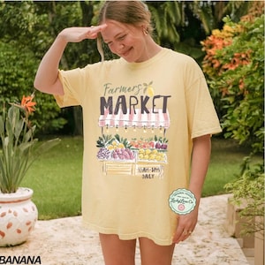 May include: A light yellow t-shirt with a graphic of a farmer's market stand, displaying various fruits and vegetables. The text "Farmers Market" is above the stand, with the hours "10AM-5PM DAILY" below. A circular logo is on the lower right.