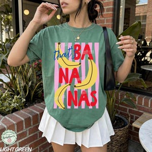 Puede incluir: Camiseta verde claro con un dise&ntilde;o gr&aacute;fico que presenta pl&aacute;tanos amarillos y las palabras "that's bananas" en una fuente l&uacute;dica. El dise&ntilde;o incluye rayas verticales rosas y texto rojo. La camiseta est&aacute; etiquetada como "Light Green".