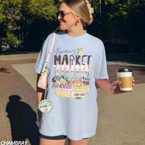 May include: Light blue t-shirt with a "Farmers Market" graphic. The design features a market stall with fruits and vegetables. The shirt also has the text "10AM-5PM DAILY". A coffee cup is held in the hand.