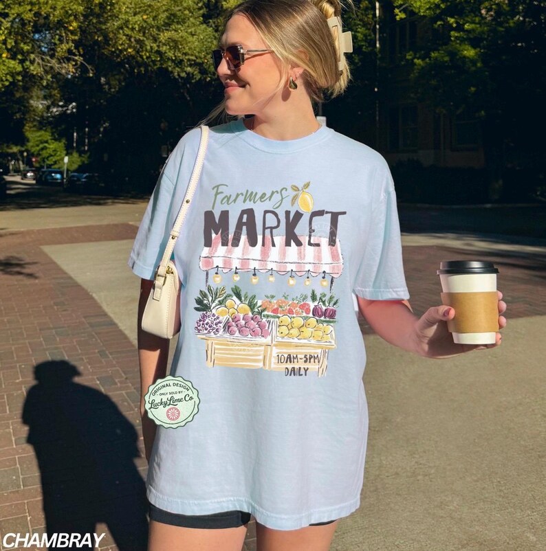 May include: Light blue t-shirt with a "Farmers Market" graphic. The design features a market stall with fruits and vegetables. The shirt also has the text "10AM-5PM DAILY". A coffee cup is held in the hand.
