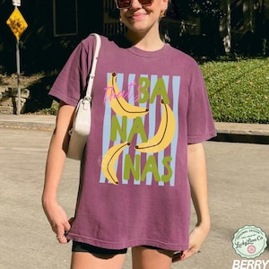 May include: Berry-colored t-shirt with a graphic design featuring yellow bananas and the text "That's BANANAS" in a playful font. The design includes blue vertical stripes and green text. The shirt is a casual, unisex style.