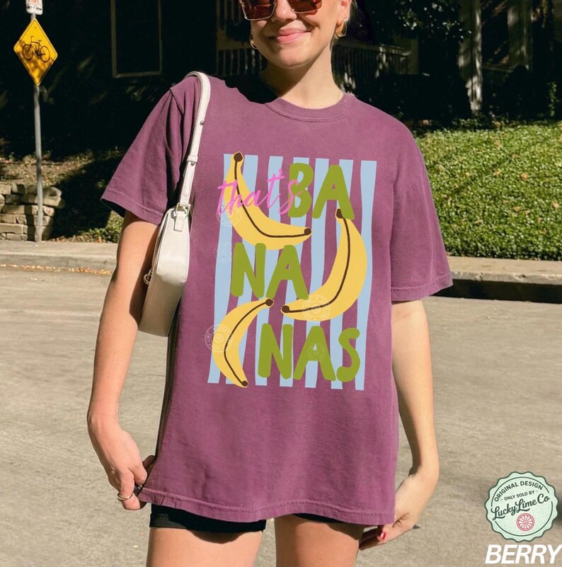 May include: Berry-colored t-shirt with a graphic design featuring yellow bananas and the text "That's BANANAS" in a playful font. The design includes blue vertical stripes and green text. The shirt is a casual, unisex style.