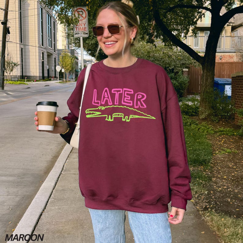May include: A maroon sweatshirt featuring the word "LATER" in pink and a green alligator design. The sweatshirt is paired with light blue jeans. A person is holding a coffee cup.