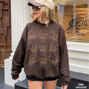 May include: A dark chocolate brown long-sleeve sweatshirt featuring a grid of nine light-colored line art skull illustrations arranged in three rows of three. The garment has a crew neckline, long sleeves, and a relaxed fit, with the text DARK CHOCOLATE.