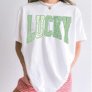 May include: White t-shirt with the word "LUCKY" in green, featuring various patterns like checkered, floral, and striped designs. The shirt is paired with red and white checkered shorts. The word "WHITE" is printed at the bottom.