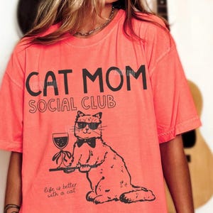 K&ouml;nnte beinhalten: Ein lachsfarbenes T-Shirt mit schwarzem Text, der CAT MOM SOCIAL CLUB und Life is better with a cat lautet. Die Grafik zeigt eine Katze mit Sonnenbrille und Fliege, die ein Weinglas auf einem Tablett h&auml;lt, dargestellt im schwarzen Linienstil.