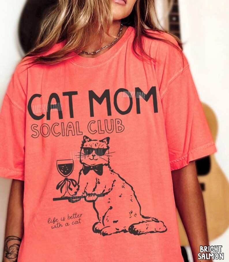 K&ouml;nnte beinhalten: Ein lachsfarbenes T-Shirt mit schwarzem Text, der CAT MOM SOCIAL CLUB und Life is better with a cat lautet. Die Grafik zeigt eine Katze mit Sonnenbrille und Fliege, die ein Weinglas auf einem Tablett h&auml;lt, dargestellt im schwarzen Linienstil.