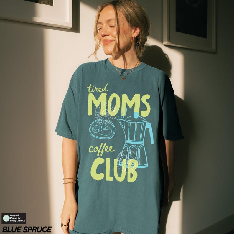 May include: Blue Spruce t-shirt with the text "tired MOMS coffee CLUB" in lime green and blue. The graphic includes a coffee pot and a pastry. The shirt is a relaxed fit.