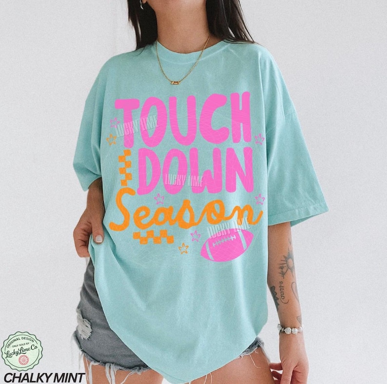May include: A light blue t-shirt with the words "TOUCH DOWN SEASON" in pink and orange. A pink football graphic is below the text. The shirt is a relaxed fit.