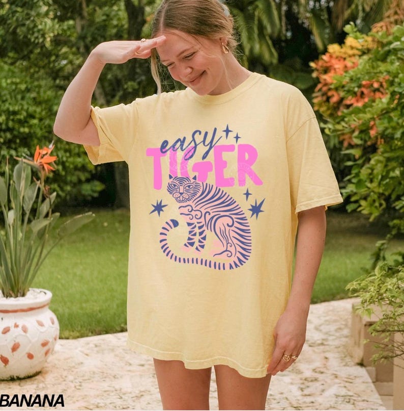 Puede incluir: Una camiseta amarilla con el texto "easy Tiger" en rosa y azul, con una ilustraci&oacute;n de un tigre. La camiseta es de corte holgado y tiene mangas cortas. El dise&ntilde;o incluye estrellas alrededor del tigre.