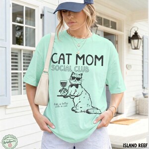 K&ouml;nnte beinhalten: Ein helles mintgr&uuml;nes T-Shirt mit kurzen &Auml;rmeln und einem schwarzen Aufdruck einer Katze mit Sonnenbrille und Fliege, die ein Weinglas h&auml;lt. Der Text lautet CAT MOM SOCIAL CLUB und Life is better with a cat. Es hat einen Rundhalsausschnitt.