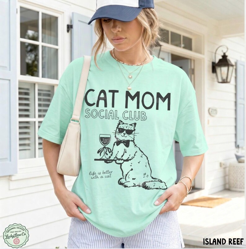 K&ouml;nnte beinhalten: Ein helles mintgr&uuml;nes T-Shirt mit kurzen &Auml;rmeln und einem schwarzen Aufdruck einer Katze mit Sonnenbrille und Fliege, die ein Weinglas h&auml;lt. Der Text lautet CAT MOM SOCIAL CLUB und Life is better with a cat. Es hat einen Rundhalsausschnitt.