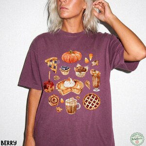 May include: A dark berry-colored short-sleeve t-shirt featuring a watercolor-style graphic print of various autumn-themed treats, including a pumpkin, a slice of pie, a caramel apple, a cinnamon roll, and mugs of warm beverages with whipped cream.