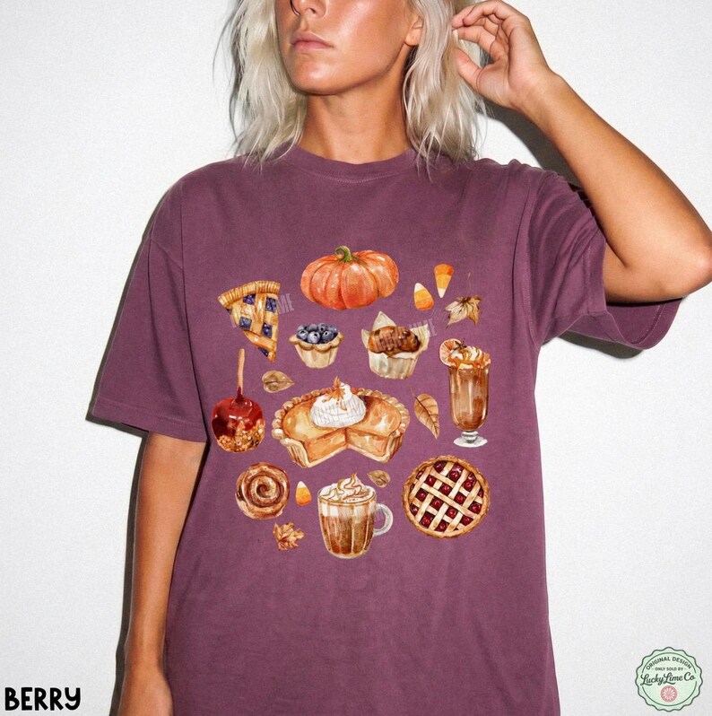 May include: A dark berry-colored short-sleeve t-shirt featuring a watercolor-style graphic print of various autumn-themed treats, including a pumpkin, a slice of pie, a caramel apple, a cinnamon roll, and mugs of warm beverages with whipped cream.