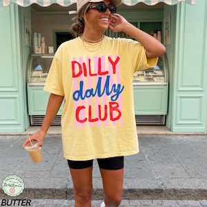 May include: Yellow t-shirt with the text "DILLY DALLY CLUB" in red, blue, and pink. The shirt is worn with black shorts. The image also includes a coffee drink in a clear plastic cup.