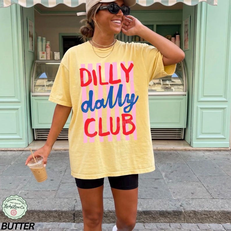 May include: Yellow t-shirt with the text "DILLY DALLY CLUB" in red, blue, and pink. The shirt is worn with black shorts. The image also includes a coffee drink in a clear plastic cup.