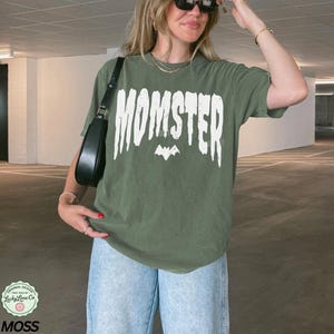 May include: An olive green t-shirt with the word "MOMSTER" in white block letters and a bat graphic. The shirt is paired with light blue jeans and a black shoulder bag. The shirt is from Lucky Lime Co.