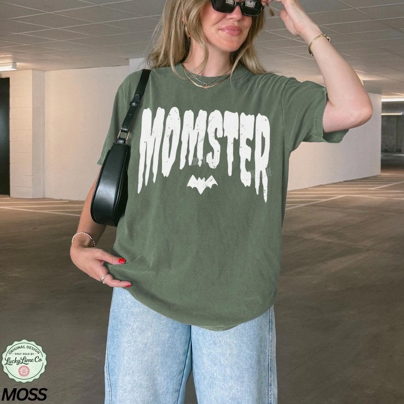 May include: An olive green t-shirt with the word "MOMSTER" in white block letters and a bat graphic. The shirt is paired with light blue jeans and a black shoulder bag. The shirt is from Lucky Lime Co.