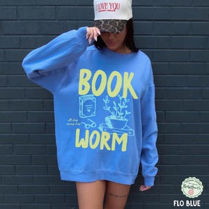 May include: A blue sweatshirt with the words "BOOK WORM" in yellow, along with illustrations of a book, glasses, and a potted plant. The text "all day every day" is also included. A white hat with the text "I love you so much" is also visible.