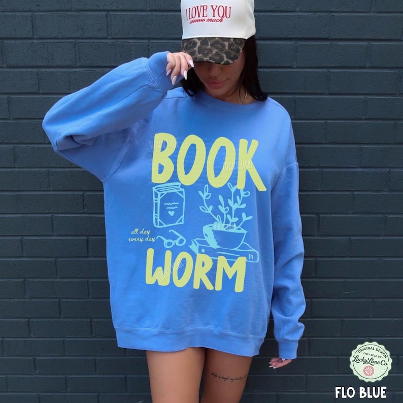 May include: A blue sweatshirt with the words "BOOK WORM" in yellow, along with illustrations of a book, glasses, and a potted plant. The text "all day every day" is also included. A white hat with the text "I love you so much" is also visible.