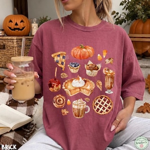 May include: A maroon short-sleeve t-shirt featuring a watercolor-style graphic print of various autumn-themed treats, including a pumpkin, a slice of pie, a caramel apple, a muffin, a cinnamon roll, a latte, and a lattice-topped berry pie.