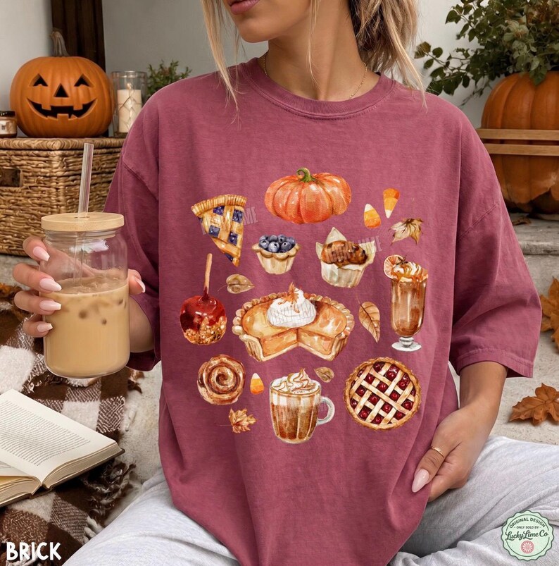 May include: A maroon short-sleeve t-shirt featuring a watercolor-style graphic print of various autumn-themed treats, including a pumpkin, a slice of pie, a caramel apple, a muffin, a cinnamon roll, a latte, and a lattice-topped berry pie.