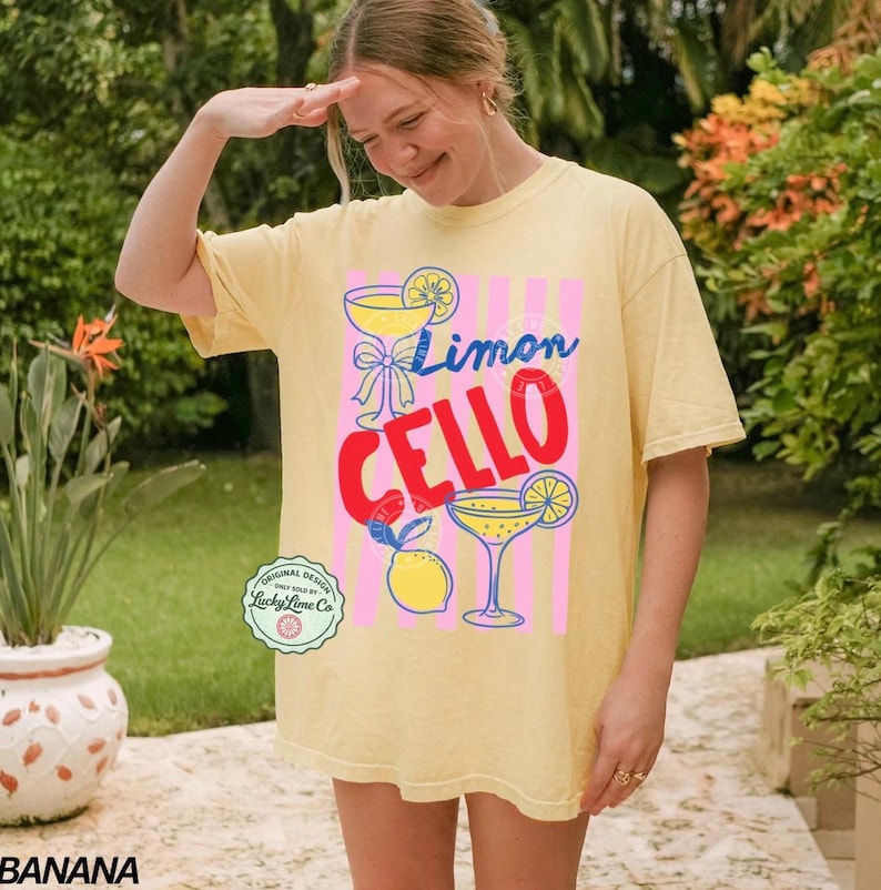 May include: A light yellow t-shirt with the words "Limon Cello" in blue and red, along with illustrations of lemons and cocktails. The shirt has a relaxed fit and short sleeves. A small circular logo is visible on the bottom left.