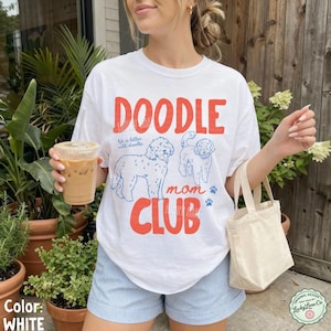 Puede incluir: Camiseta blanca con las palabras "DOODLE mom CLUB" en rojo y azul. La camiseta presenta ilustraciones de dos doodles y la frase "life is better with doodles". La camiseta se usa con pantalones cortos a rayas azules y blancas.