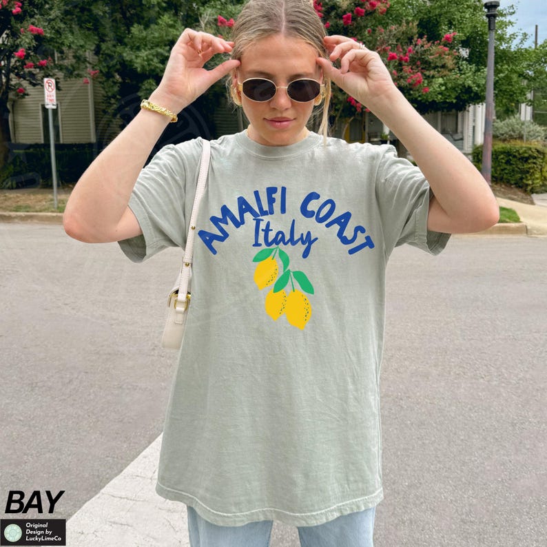 K&ouml;nnte beinhalten: Ein hellgr&uuml;nes T-Shirt mit einer blauen und wei&szlig;en Grafik, auf der "Amalfi Coast Italy" mit einer Gruppe gelber Zitronen steht.