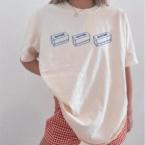 Puede incluir: Una camiseta blanca de gran tama&ntilde;o con un gr&aacute;fico de tres envases de mantequilla en barra con la palabra "BUTTER" impresa en cada uno. La camiseta la lleva puesta una persona que lleva pantalones cortos de cuadros rojos y blancos.